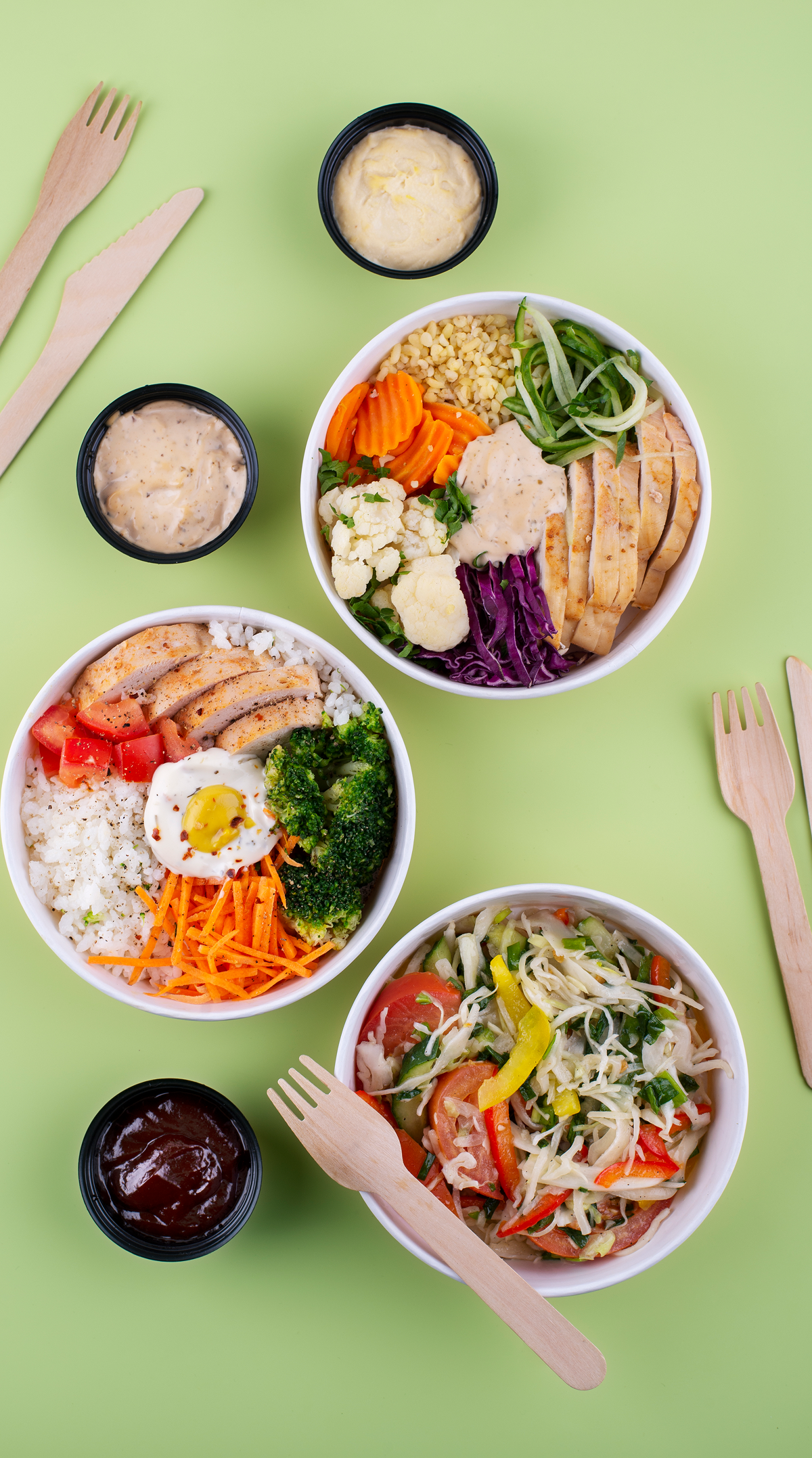 Bowls of colorful healthy bowls with rice, vegetables, and sauce, on a green background with wooden forks nearby.