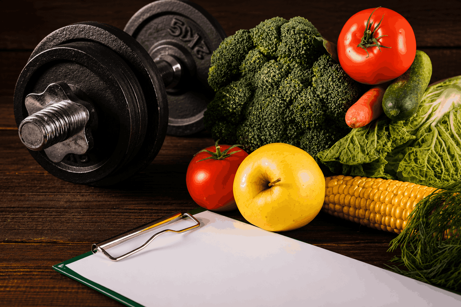 Dumbbell weights sit on a dark wood table beside fresh vegetables and an apple, with a clipboard in the foreground. (Healthy living scene)