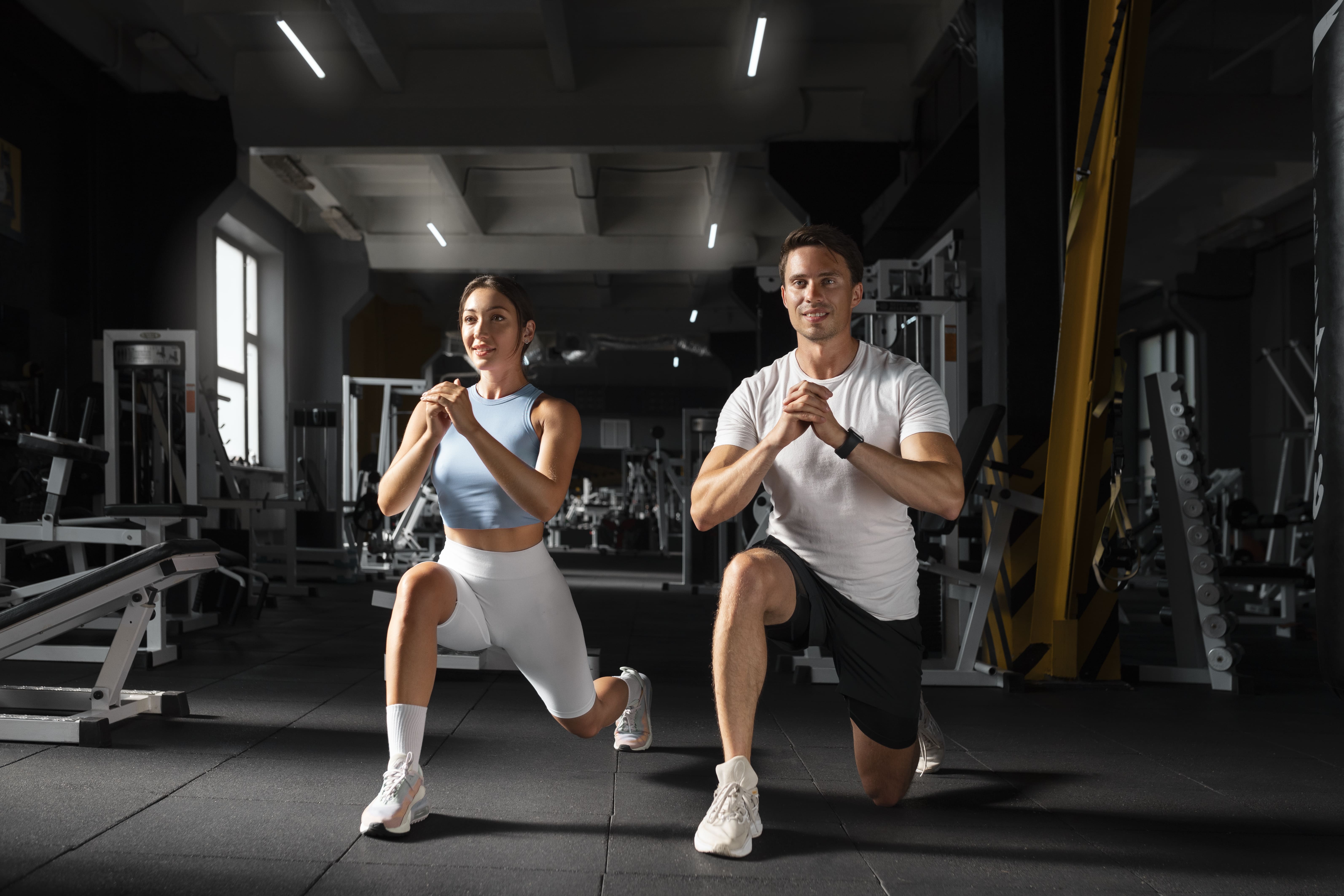 woman-helping-man-gym 1 - mistalife Two people perform synchronized lunges in a gym, facing forward with determined expressions.