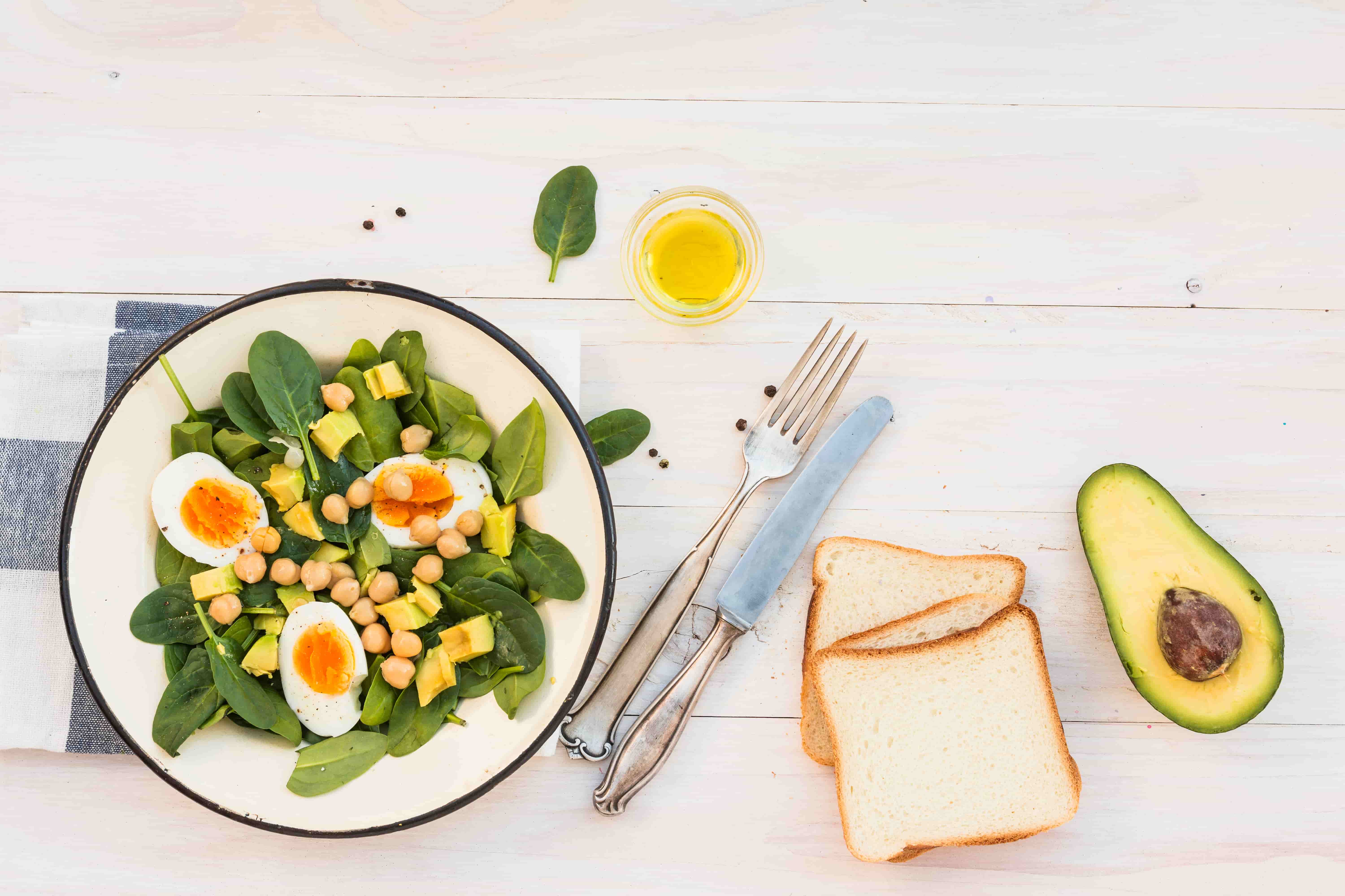 Healthy Meals for Weight Loss 2 - mistalife Salad on a white plate with spinach, avocado, chickpeas, cubed cheese and halved boiled eggs on a light wooden table near bread slices and cutlery.