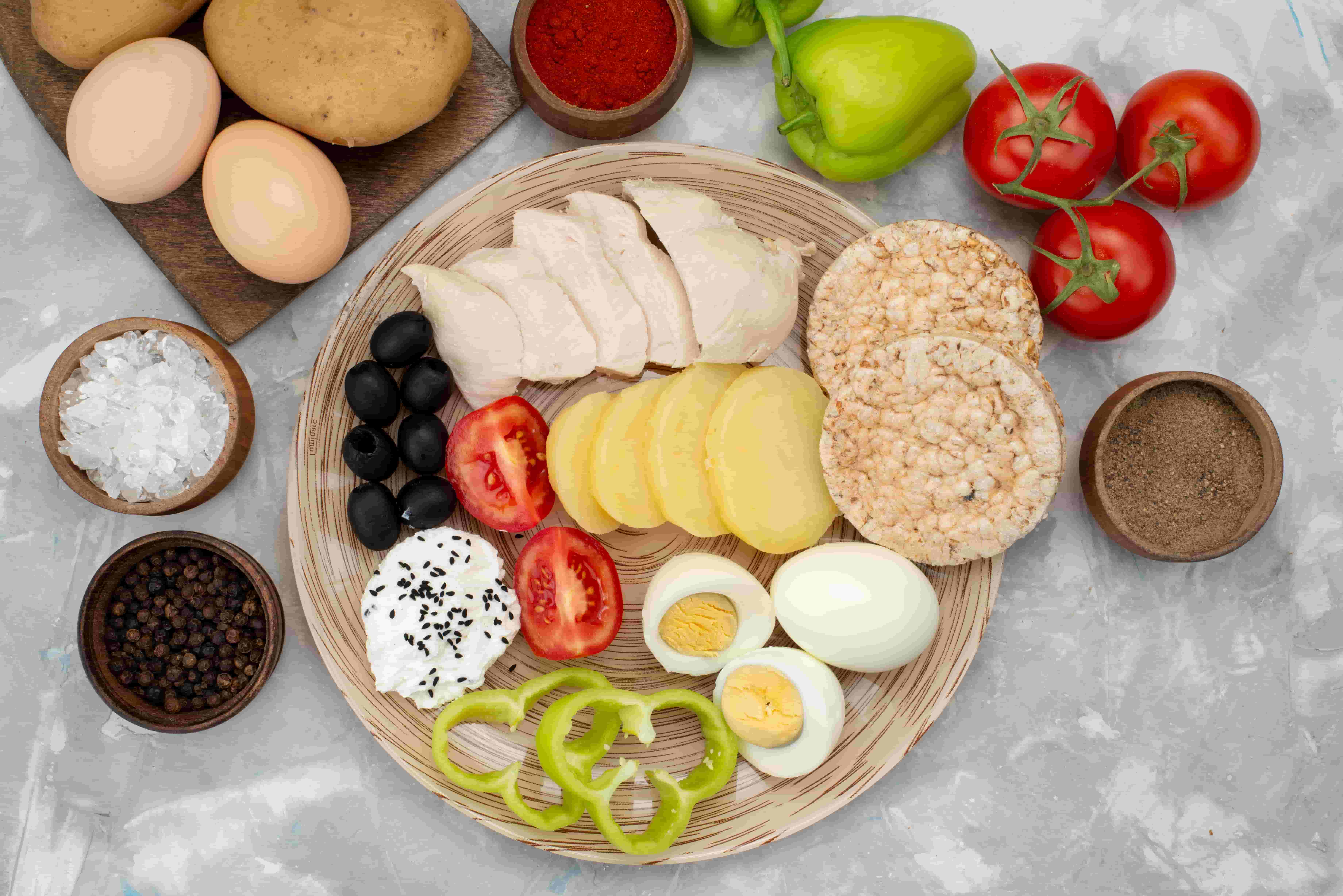High-Protein Diet for Weight Loss - mistalife A round wooden board holds sliced turkey or chicken with cheese slices, black olives, tomato slices, hard-boiled eggs, and green bell pepper rings, surrounded by crackers and vegetables on a gray surface.